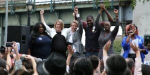 A group of 2 white women, a black man and black woman hold their fists in the air on the podium at a rally