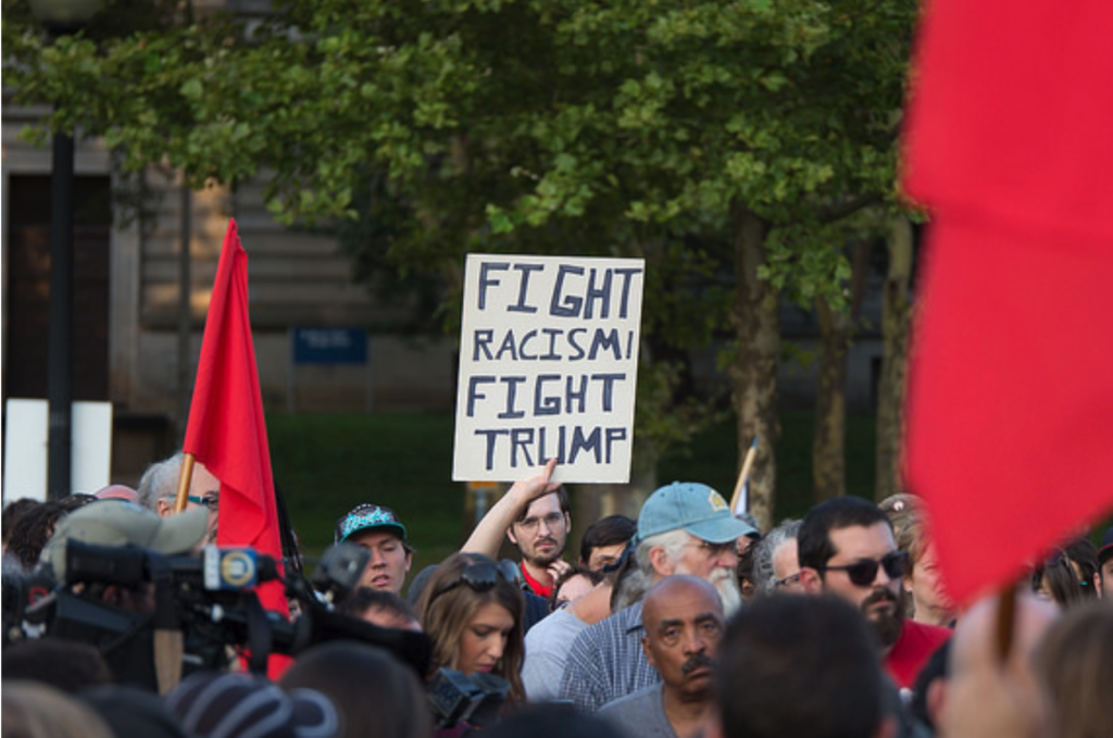 Photo of people at a protest with one person holding up a sign that says Fight Racism Fight Trump