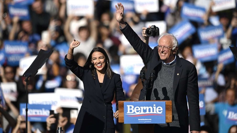 Alexandra Ocasio-Cortez and Bernie Sanders at a rally, holding their fists up in the air