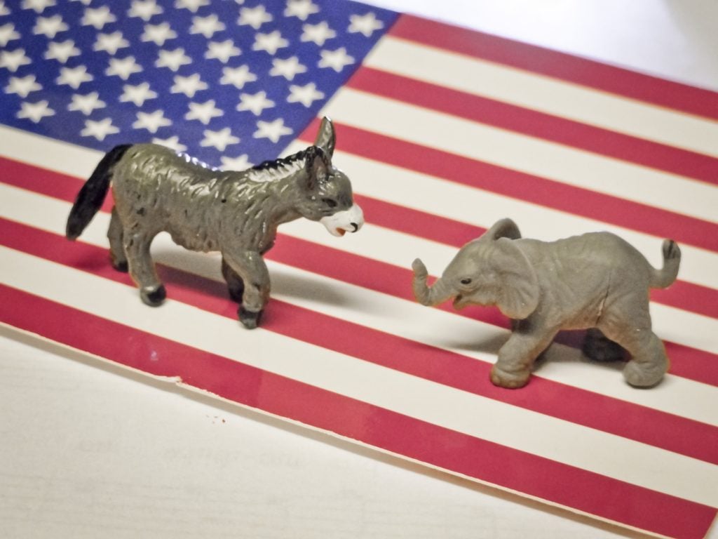 Photograph that shows an american flag with a donkey figurine and an elephant figurine