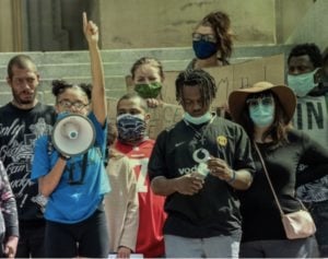 Knot of people protesting in front of steps leading into a civic building. A slim woman in a bright blue T-shirt is holding a bullhorn close to her mouth with one hand, pointing straight up with the other. People are masked, various skin hues from deep black to olive.