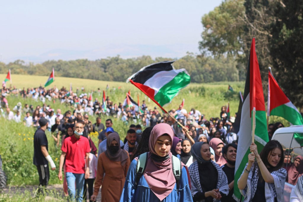 Photo of Palestinians marching through the ruins of the village of Al-Lujjan, destroyed in the Nakba of 1948. Women lead the march carrying Palestinian flags.