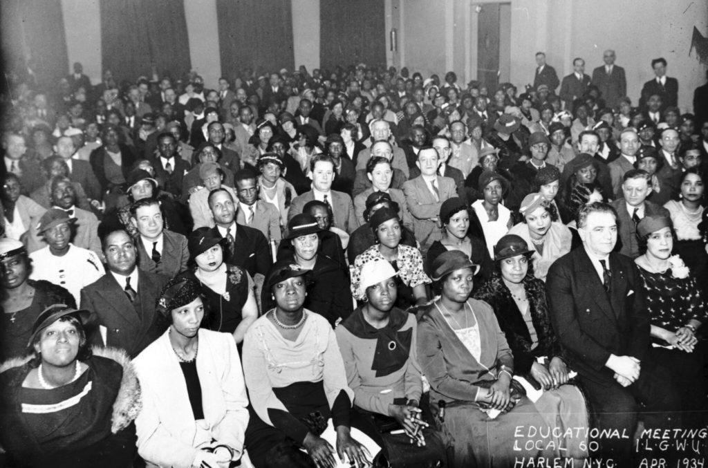 Photo from the 1930s, room full of people sitting in rows of chairs. It’s a multi-racial crowd, about half women. Everyone is nicely dressed, the women with hats, the men in suits and ties.
