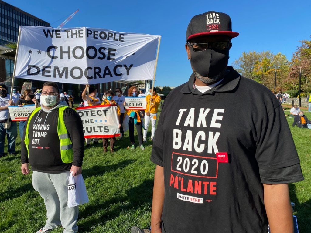 A man and a woman standing in an open, grassy area wearing dark gray shirts with red and white lettering :Take Back 2020, Pa’Lante! A small crowd is gathered several feet behind them. At the front of the crowd, two people holding up a long horizontal white banner with black lettering that reads “We the people choose democracy.”