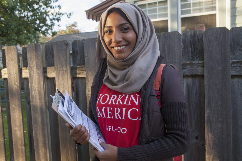 A woman with a warm smile stands in front of a wooden fence holding a clipboard. She wears a red T-shirt that says “Working America AFL-CIO,” and a beige hijab.