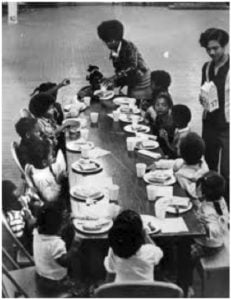 Children sitting on either side of a long table eating lunch off of trays