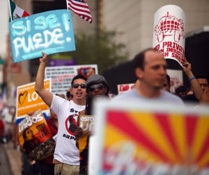 People marching in a protest..protester at the front has a sign with the Arizona flag on it