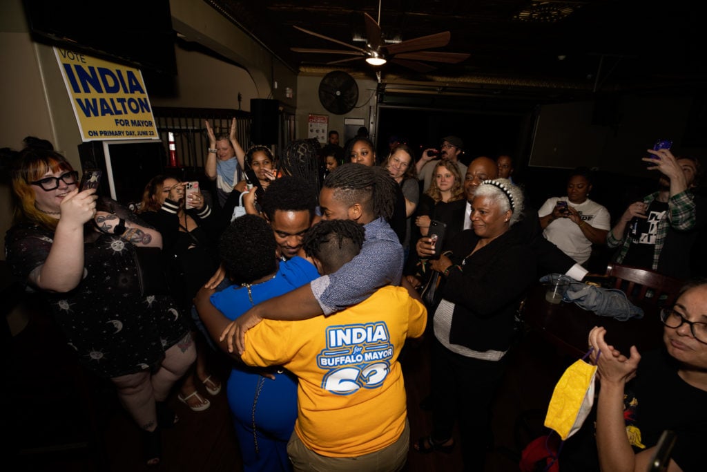 In the foreground, a Black woman in an electric-blue dress with her back to the camera, held by three young men in a group hug. The man with his back to the camera has a gold T-shirt that says “India for Buffalo Mayor.” They’re in a room with low light, with a multi-racial group of women gathered in a semi-circle around them.