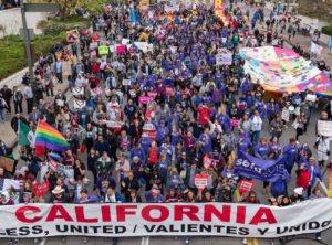 Mass demonstration, shot from above. Bright T-shirts, lots of red and purple. Lead banner reads, in English and Spanish: “California: Fearless, United/Valientes y Unidos.”