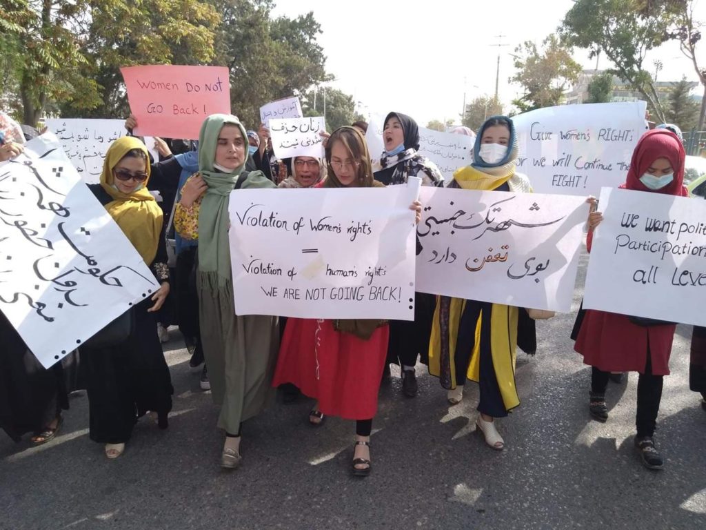A group of Afghan women in loose flowing dresses and hijabs holding signs handwritten on white poster board with black markers, some in English and some in a flowing foreign script.