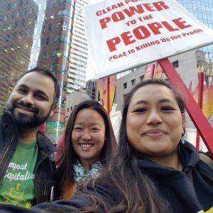 Close-up of a man and two women, one holding a sign that says “Clean Power to the People: PG&E is killing us for profit,” with steel-and-glass skyscrapers in the background. All are people of color, ethnicity ambiguous, possibly Latino (the man) and AAPI (the women).