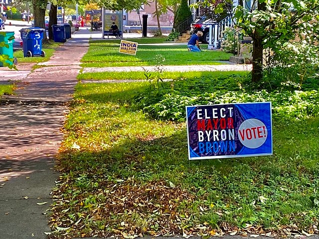 Political signs on green lawns on a tree-lined street. “Elect Mayor Byron Brown” in the foreground, “India Walton” in the background.