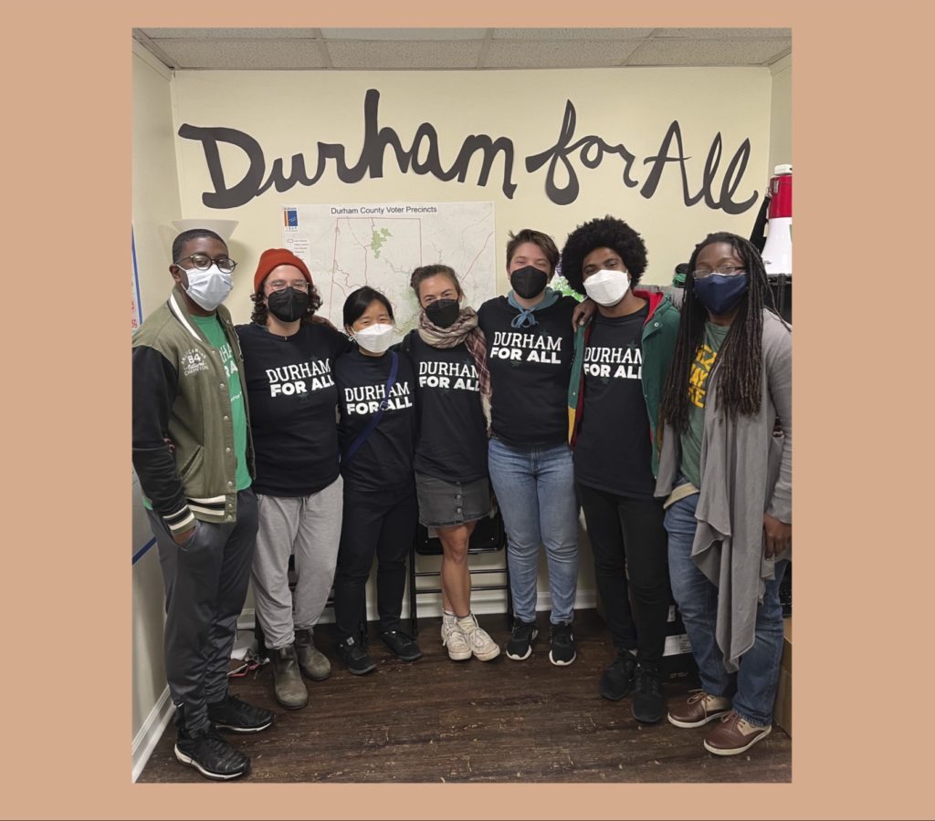 Diverse group of young people stand in a horseshoe with arms around each others’ shoulders. They’re casually dressed in jeans and sweats. Most wear a black T-shirt printed with “Durham for All” in white letters. “Durham for All” is painted on the wall behind them.