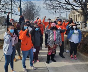 A diverse group of about 15 people posed with raised fists. They’re wearing warm sweatshirts and jackets, several of which are neon orange. Everyone is masked. In the background, bare trees and columned public buildings.