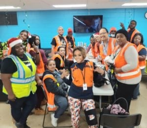 A diverse group of people, mostly women, wearing orange safety vests, gathered around a table in a break room and smiling.