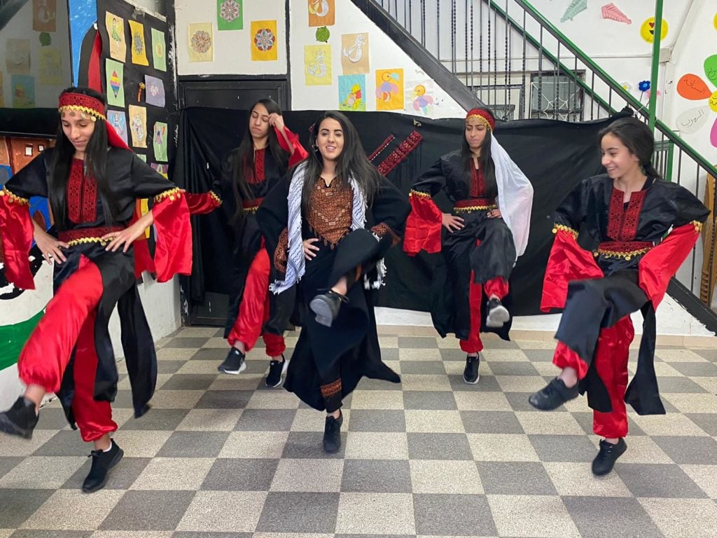 Young women dressed in black and red practicing traditional Palestinian debkeh dancing in a courtyard.