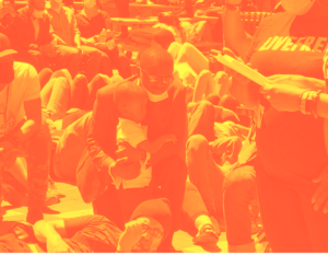 People are lying on their backs on a brick plaza, knees up, feet on the ground. In the center of the photo stands a young Black boy in a white shirt. A Black man in a neat grey suit leans over, wrapping an arm around him as if to comfort him.