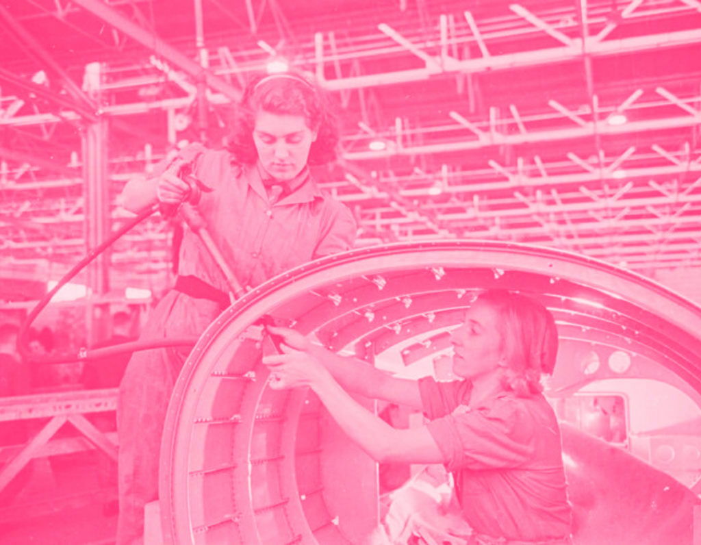 Women in 1040s clothes and hairstyles working in an industrial setting, one inside a conical piece of metal, the other above. They are building an airplane.