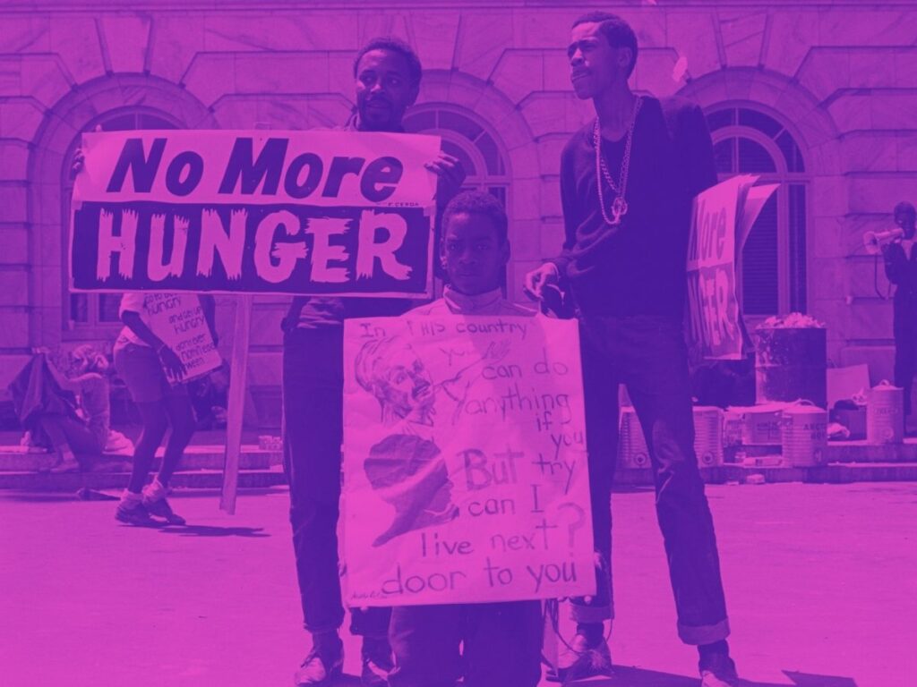 1968 photo of 3 Black demonstrators for the Poor People's Campaign. One hold a sign that reads "No More Hunger." Another holds a sign that reads "In this country you can do anything if you try, but can I live next door to you?"