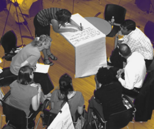 Participatory process - some people sitting in a circle, leaning in, one person writes on a long sheet of paper.