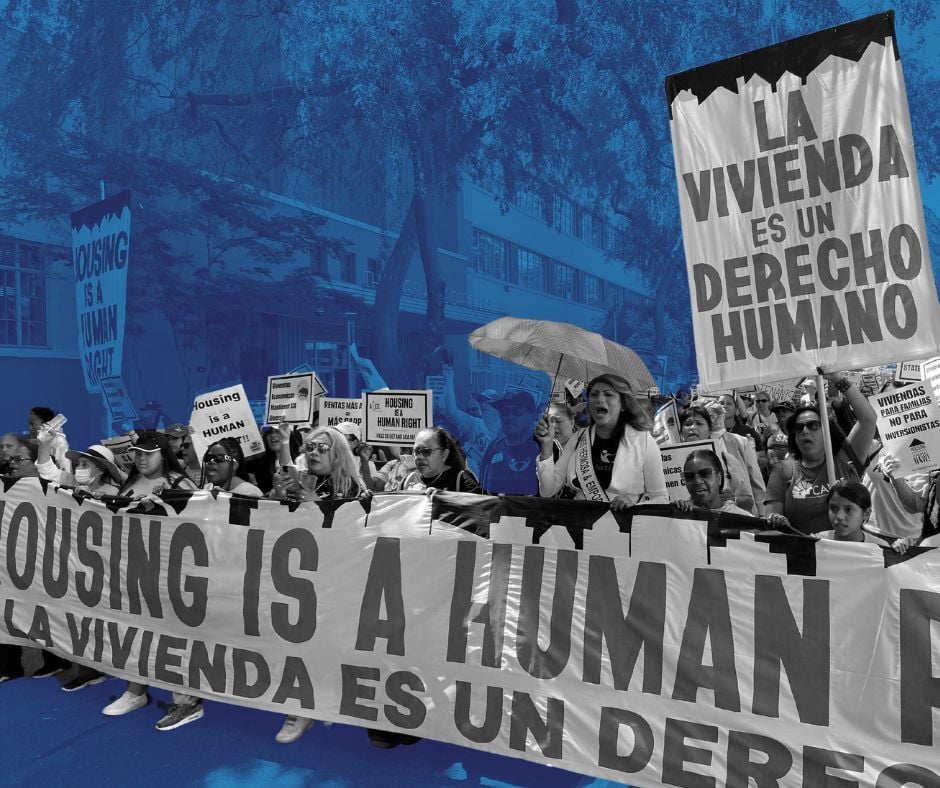 Front of a march, with people holding a long banner waist-high. It reads "Housing is a Human Right/ La Vivienda es un Derecho Humano."