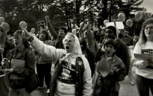 Multiracial crowd of women, many yelling, some fists up, front and center an older-looking white woman in a kerchief and a sweatshirt that reads, "i Don't Want to Strike."
