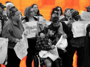 Diverse group of people standing, holding sheets of paper with names on them. Front and center, a young man in a wheelchair leaning into another person, being comforted.