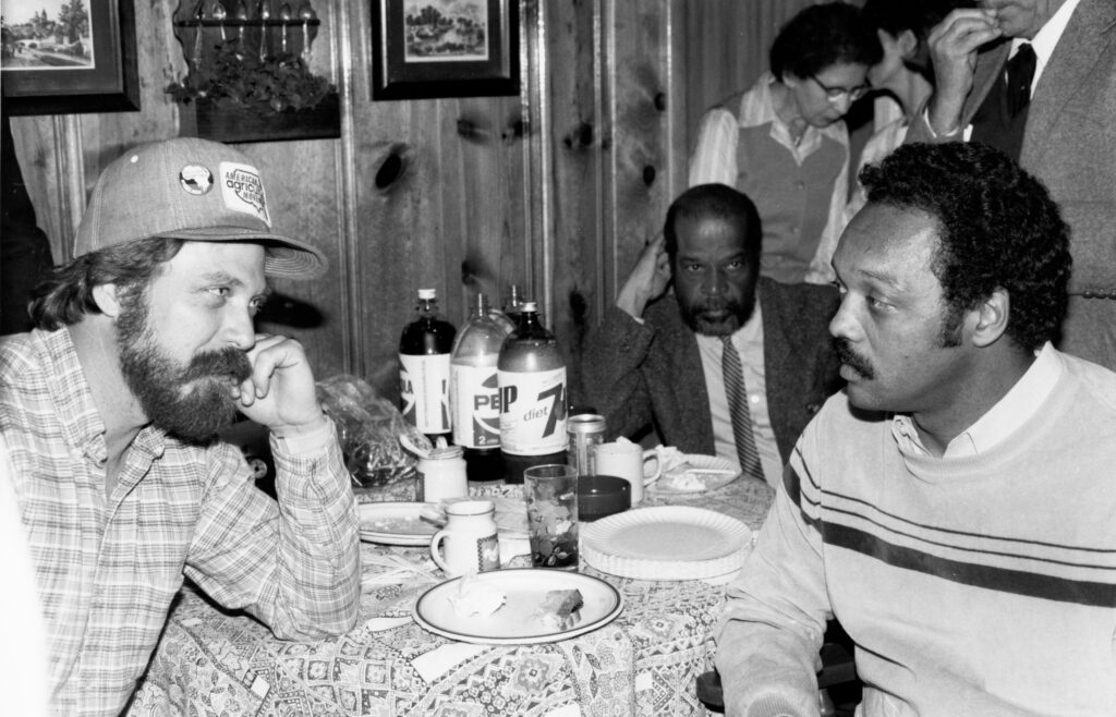 Rev. Jesse Jackson at a table across from a white man in a baseball cap.
