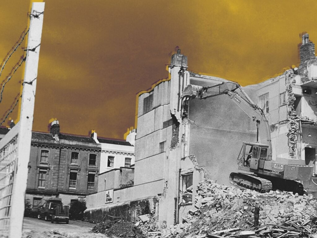 Foreground, a four-story flat-roofed apartment building being demolished, qith a bulldozer sitting on a pile of rubble. Background, a lower buidling still standing.
