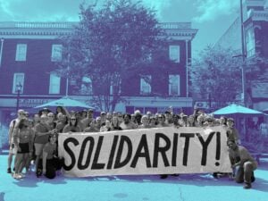 Crowd of people posed with a long banner that reads "Solidarity." They're standing in front of a flat-roofed brick building, wearing T-shirts and shorts or pants. Crowd includes both men and women. Most appear to be white but a few are not.