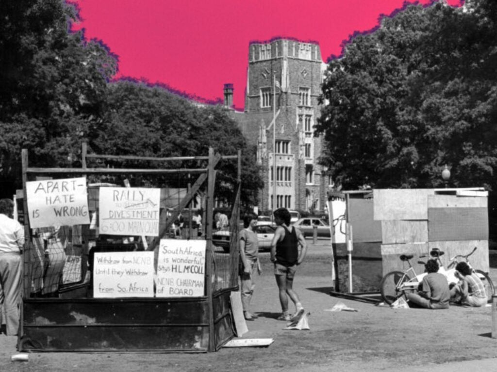 Structure made of cardboard and sticks, with signs denouncing apartheid attached, on a grassy area with an old elegant stone college building in the back. A few people are standing and sitting near the structures.