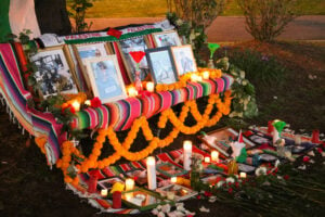 Altar with candles, photos, flowers, little Palestinian flags, on a multicolored striped Mexican blanket - main color is red.