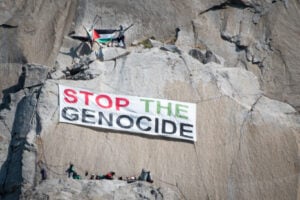 A large banner reading "stop the genocide" hanging from a sheer rock wall. Above and below it are people and climbing gear; the people above it have a Palestinian flag.