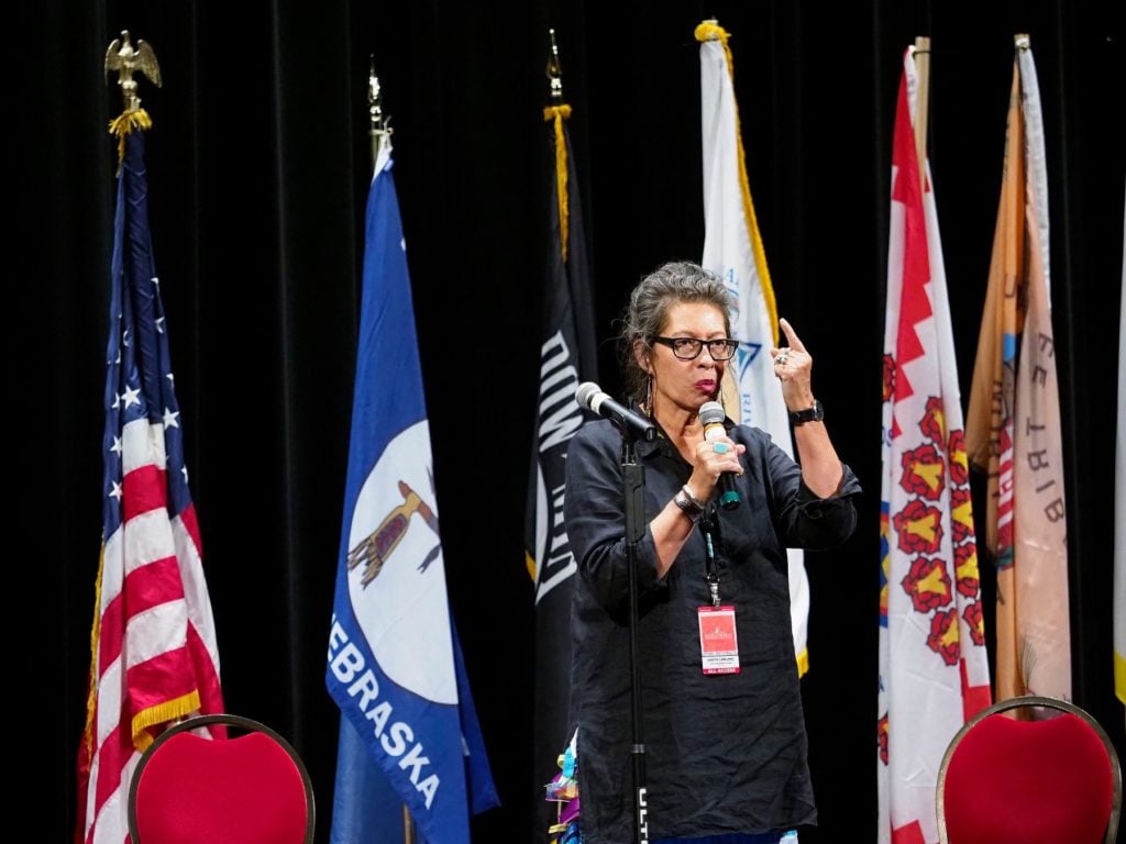 A woman with gray hair holding a mic with a variety of flags in the background