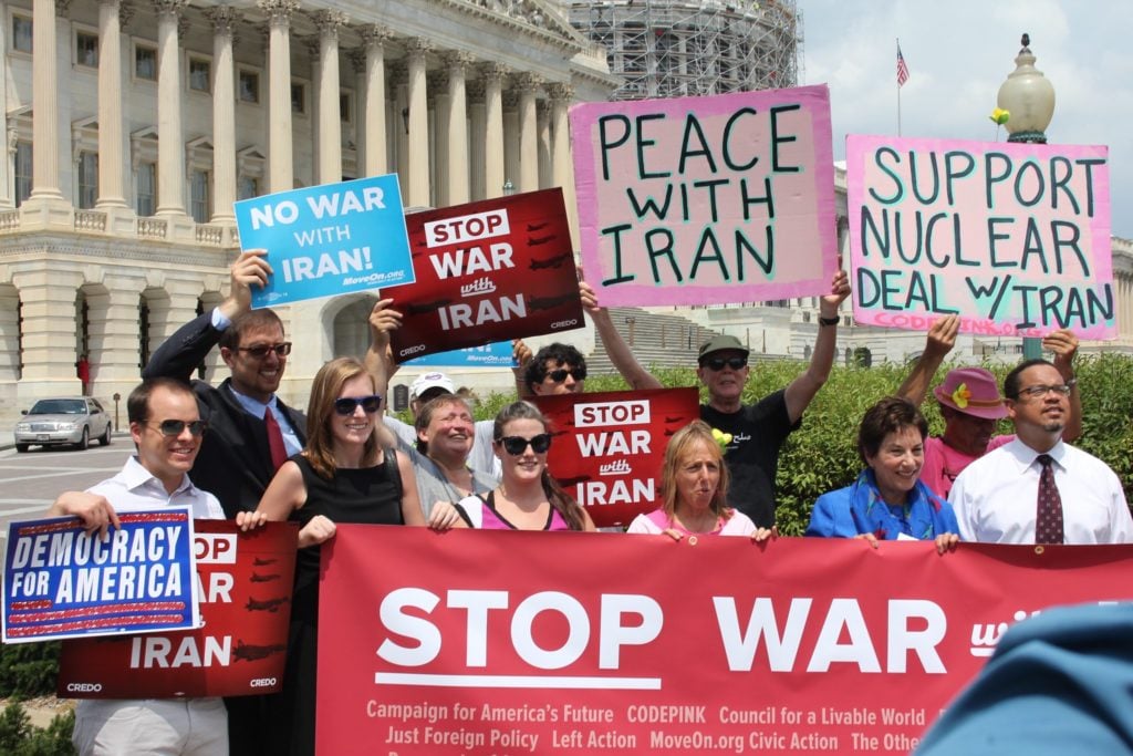 Protesters at an anti war rally holding up a variety of signs behind a big red banner that says Stop War