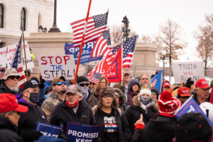 Daytime rally, white QAnon and "Stop the Steal" supporters stand maskless shoulder to shoulder outside the Minnesota State Capitol. They are carrying banners, Trump Flags and American Flags. The State Capitol is in the background.