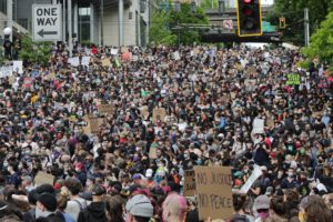 Huge crowd packing a street, seen from above and behind. A few readable signs: No Justice No Peace, Black Lives Matter. Three traffic lights at the top of the photo.