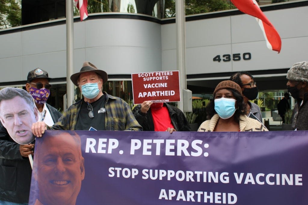A line of people in masks holding a banner that reads: “Rep. Peters: Stop Supporting Vaccine Apartheid.” They’re a diverse group: white, Black, South Asian. Man at the end of the banner holds a cardboard cutout of a face, a white man with gray hair, probably Rep. Peters.