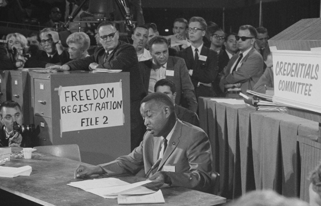 Black-and-white photo. An African American man in a white shirt, suit and tie is seated at a table reading. Several white people stand over him as he does, mostly men, some in sunglasses, expressions ranging from hostile to indifferent. A file cabinet behind the reader to the left has a hand-written sign, “Freedom Registration File 2”; a sign taped to a lectern identifies the credentials committee.