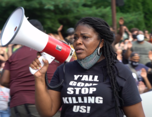 A Black woman speaks into a bullhorn. She has high cheekbones, long braids, and a serious expression. She’s wearing a black T-shirt with white letters that reads “Y’All gone stop killing us!”