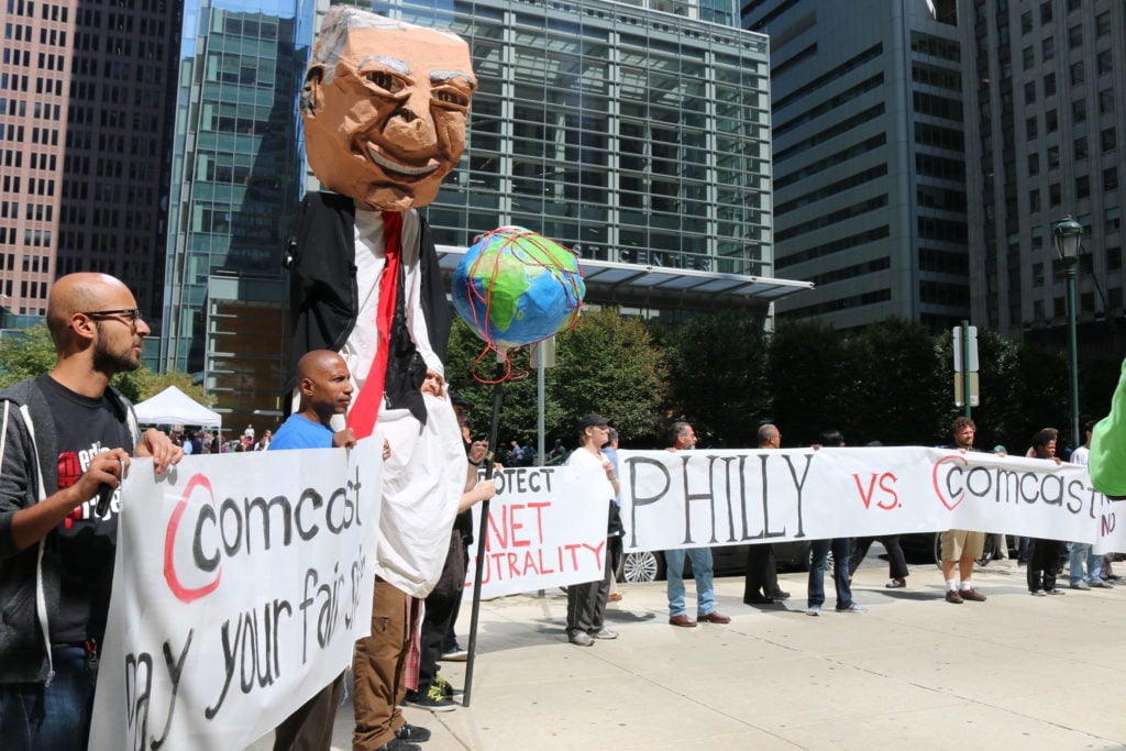 A puppet topped with a putty-colored papier mâché head of a man in glasses and a suit jacket and red tie towers over a semi-circle of people holding signs reading “Comcast pay your fair share” and “Philly vs Comcast.” A globe wrapped in red tape floats over the people, who stand in a plaza with shiny office buildings in the background.