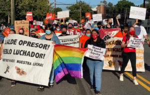 Small crowd marching down the street with banners and signs in Spanish and English, and a rainbow flag near the front. Most of the marchers are women, Latina and Black. Everyone is masked. Several have red T-shirts and signs.