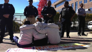 Photo shows 3 people sitting on the ground with arms wrapped around each other at a protest