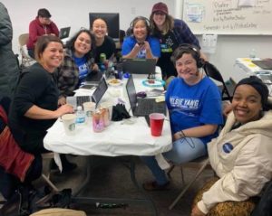 Group of women sitting on folding chairs around a table. Some have laptops; there are plastic cups and canned drinks and some snacks on the table. They’re of various ethnicities; some have cornflower blue T-shirts with “Nelsie Yang” printed in white letters.