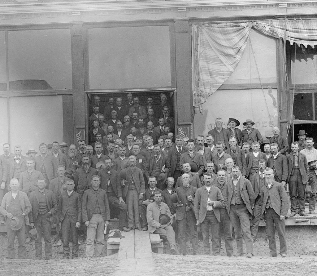 Old, partly faded black and white photo of a large group of white men posing outside a rundown meeting hall.