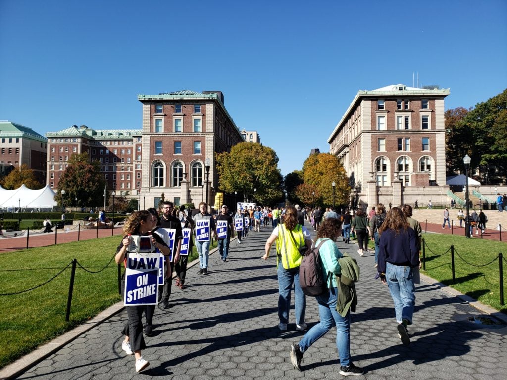 People marching in an oval along a long walkway with old brick buildings in the background. They’re carrying blue-and-white signs that read “UAW on strike.” Most appear young, are wearing blue jeans.