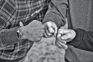 Black-and-white close-up of hands fist-bumping. One has darker skin, the other two lighter. All have rings on their first finger.