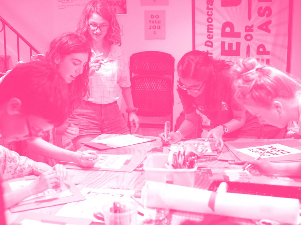 Young women lean over a table, pens and markers in hand.