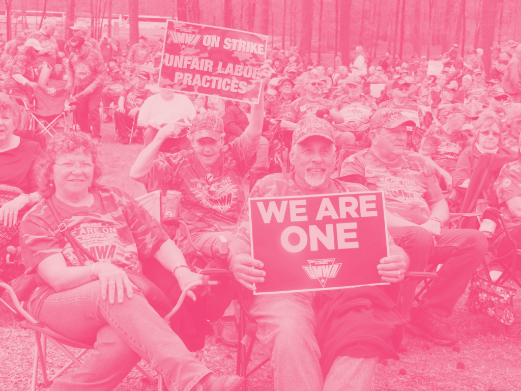 White people sitting in lawn chairs wearing UMWA T-shirts and caps; sign in the foreground reads "We Are One," and one in the background says "UMWA On Strike."