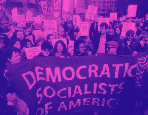In the foreground, a big red banner with white lettering that reads “Democratic Socialists of America.” People in heavy coats, scarves and hats stand behind it. Most are white. The thick crowd extends back down the city street. Lots of people are holding other signs, white with red letters, saying “Working women of the world unite.”